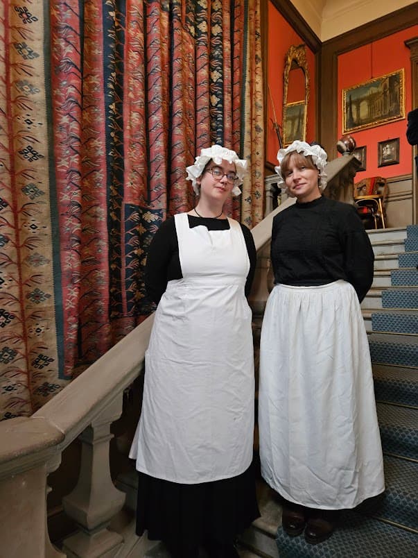Two women dressed as Edwardian house maids in long white aprons, black dresses and hat stand on the staircase at Edinburgh's Lauriston Castle