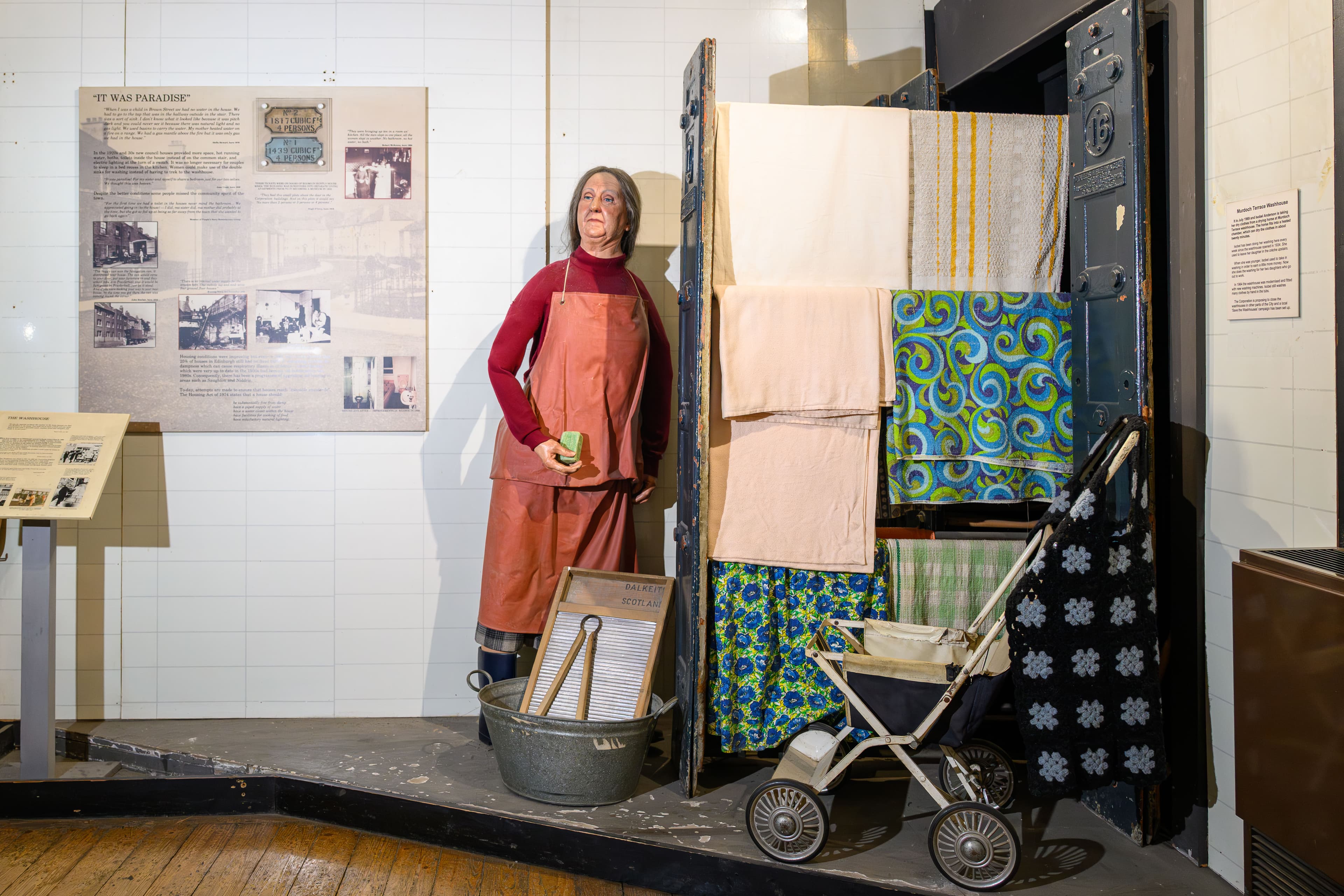 A woman is positioned next to a cabinet, dressed in period clothing, with a baby carriage nearby, in a cozy museum setting.