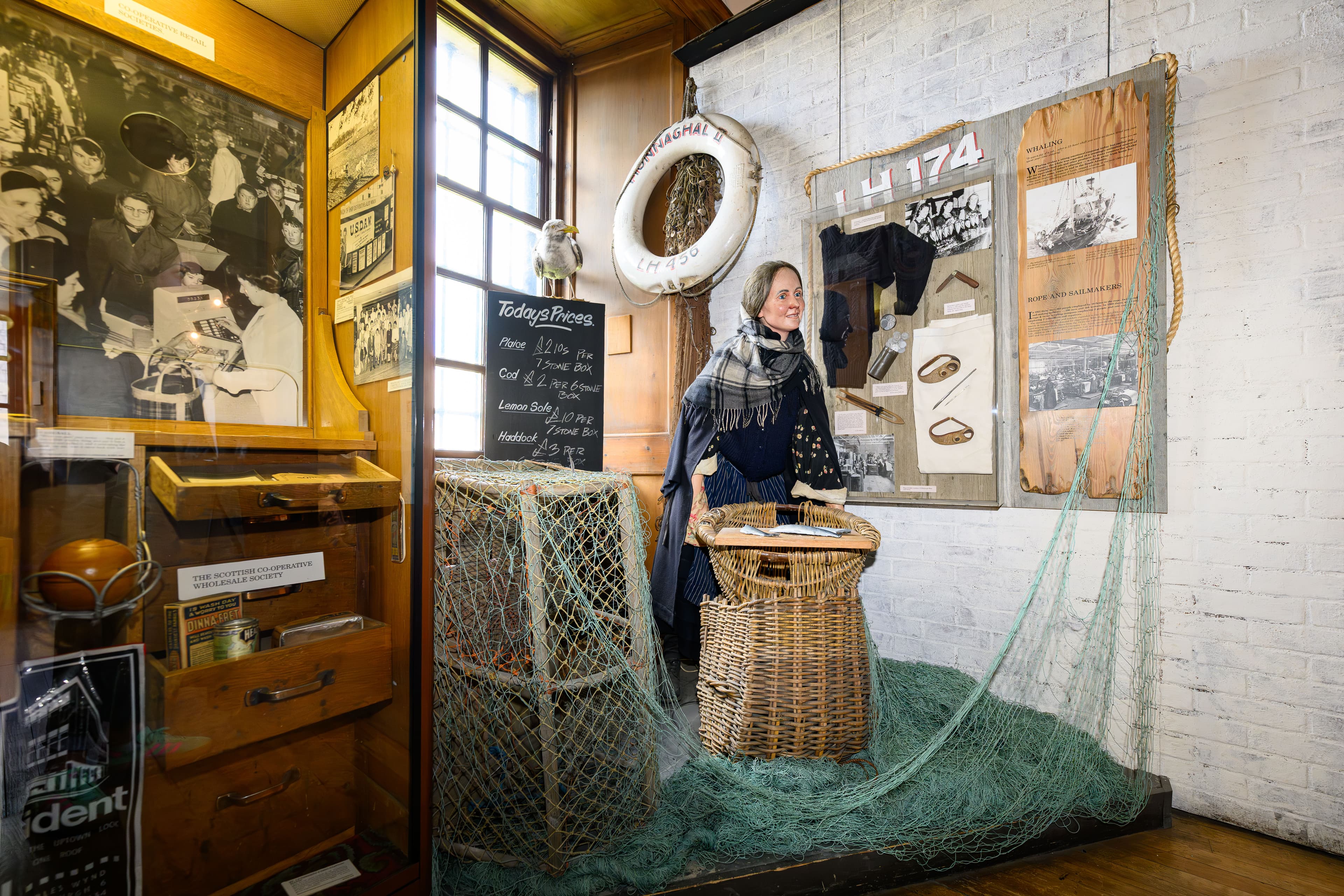 A fisherwoman in a blue dress stands beside a woven basket, smiling gently in a bright outdoor setting beside fishing nets
