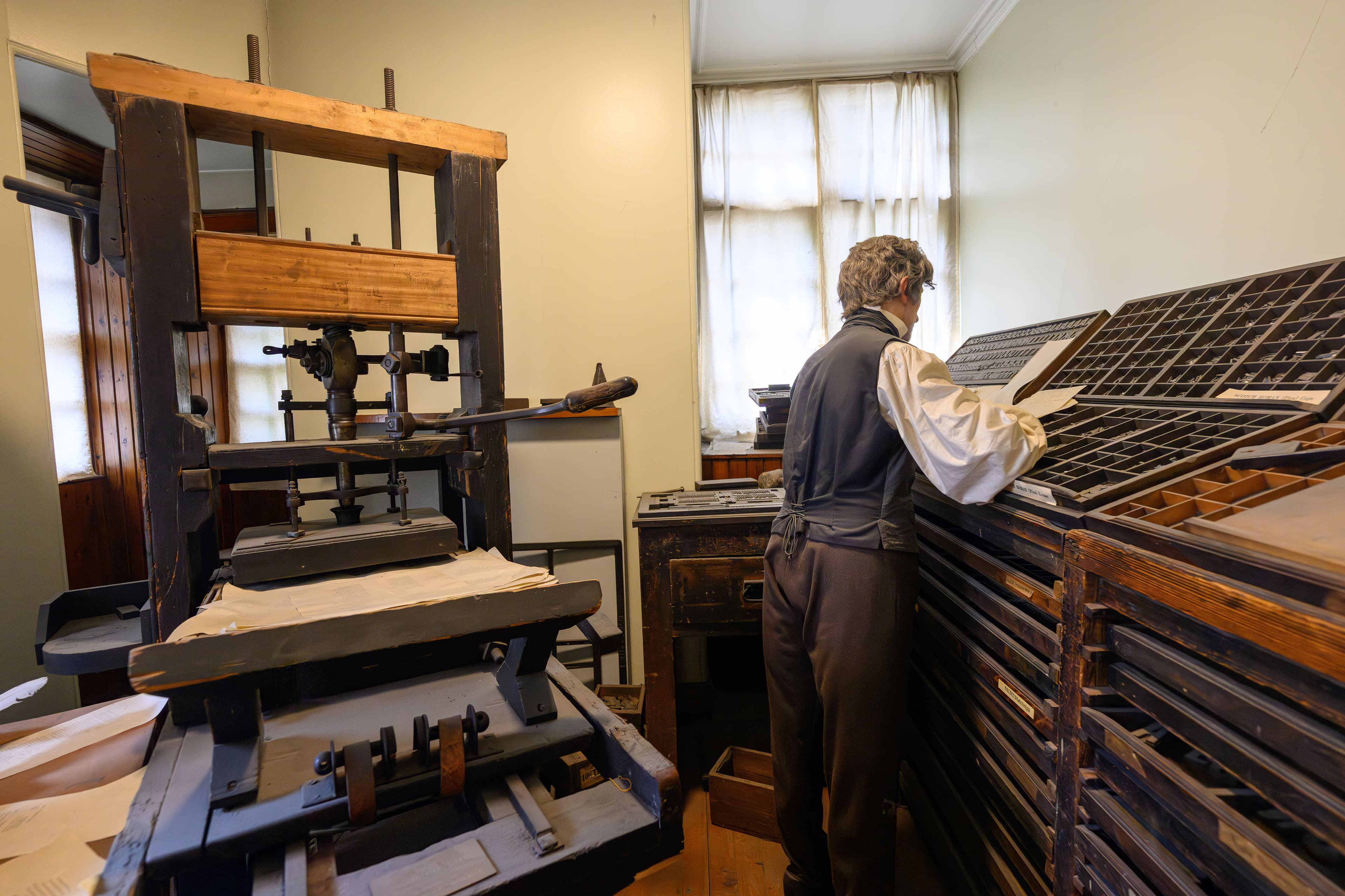 A mannequin in a shirt and waistcoat is engaged in operating a printing press, showcasing craftsmanship in a printing room environment.