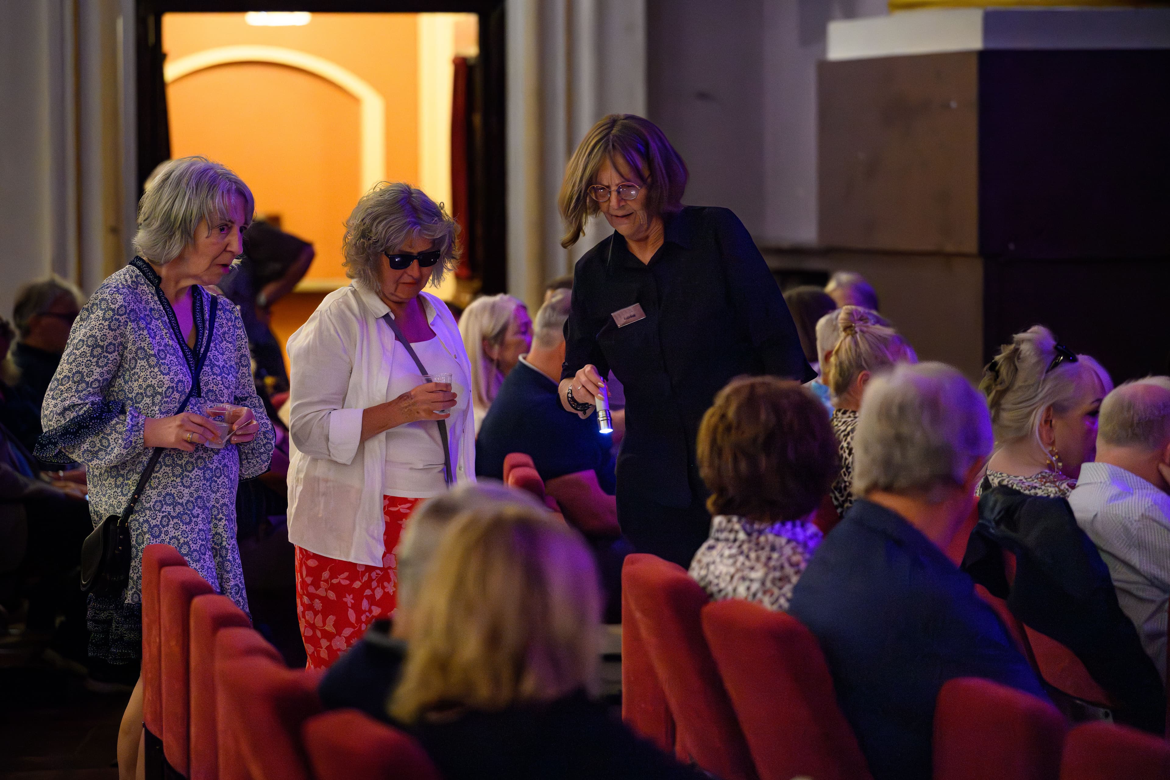 A woman converses with an front of house attendant in the stalls of the Usher Hall, both engaged in looking for the correct seat to sit in.