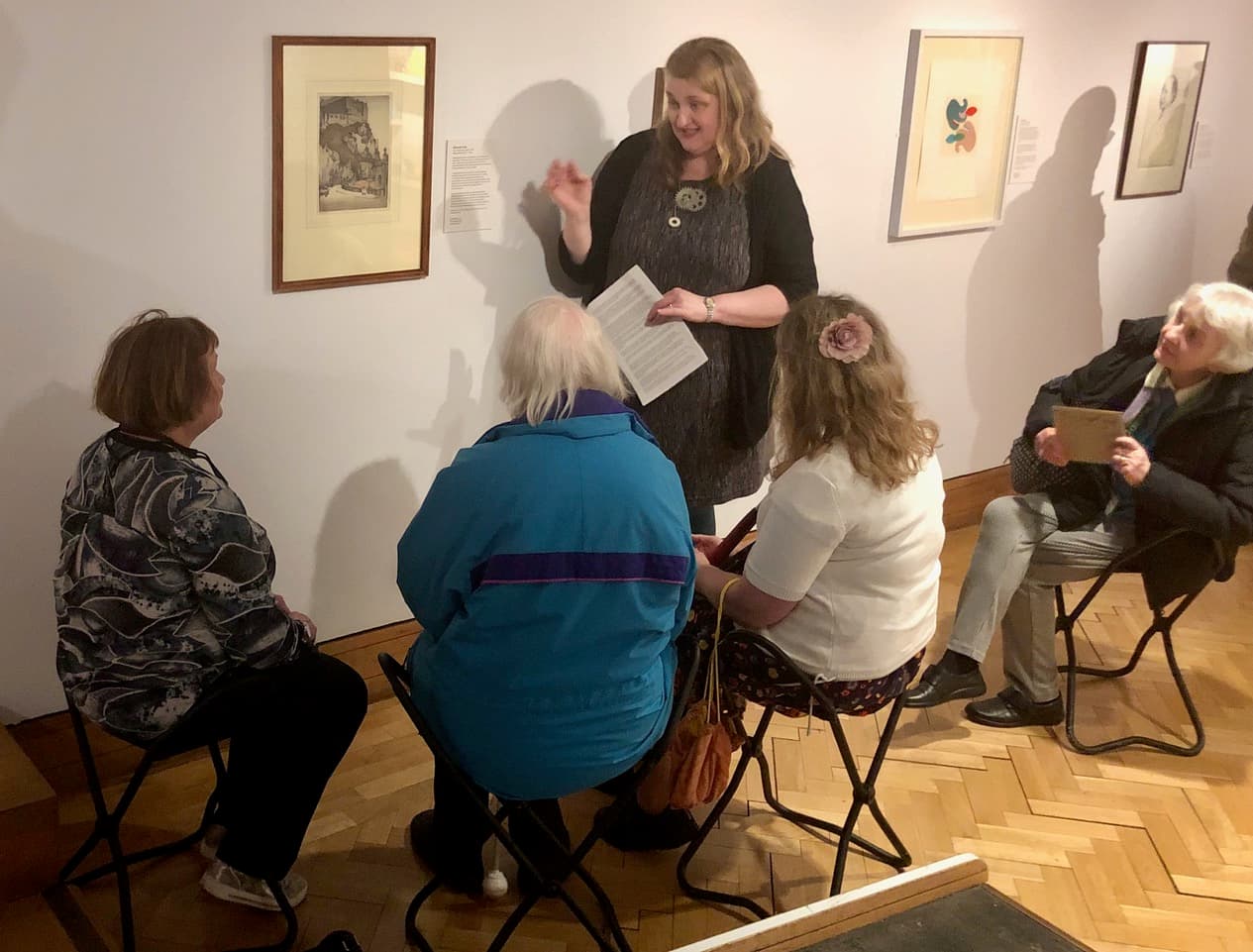 In an art gallery, a woman speaks to a small group, sharing insights about the art on display. 