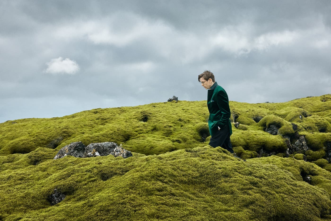 Pianist Víkingur Ólafsson wears a green jacket and is walking over a moss-covered hill. A moody sky is overhead.