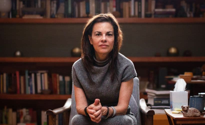 Dr Orna Guralnik, seated, dressed in grey leaning forward with hands clasped.  Large bookcase in the background
