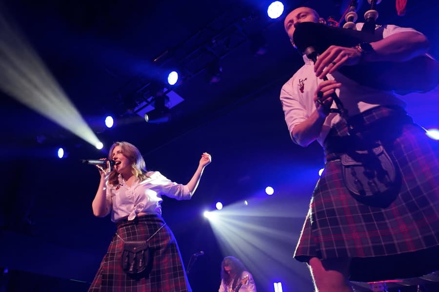 A photo of two performers in kilts - the woman singing, and the man playing bagpipes.
