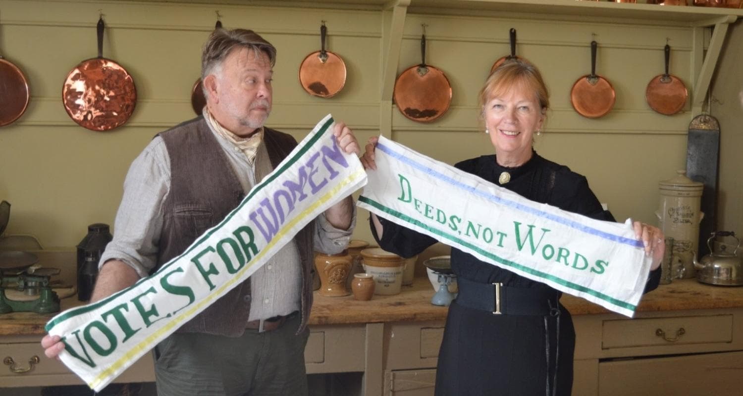 Two individuals wear Edwardian costume and hold a banner each reading 'Votes for women' and 'Deeds not words' 