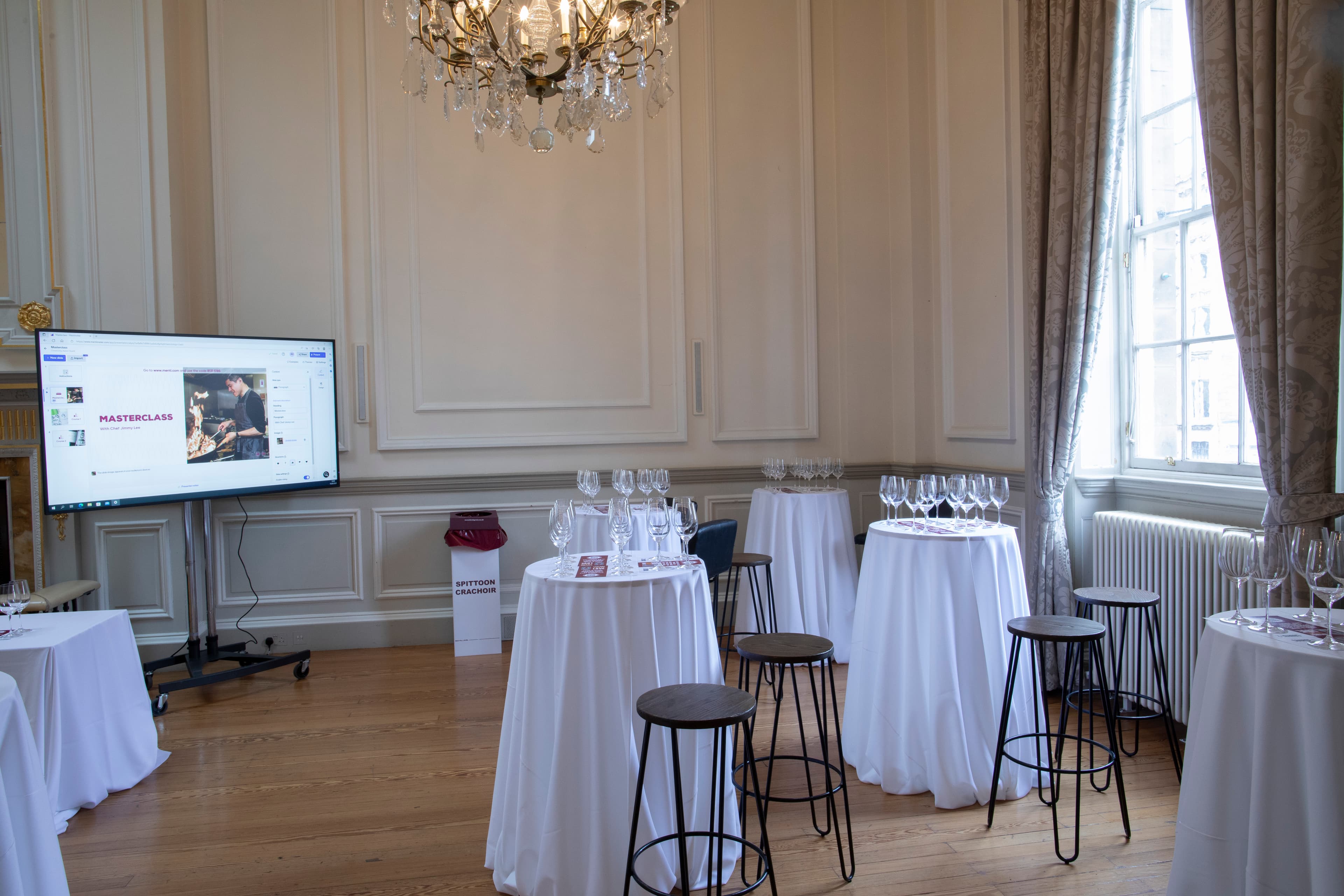 A room prepared for a party, showcasing tables with glasses on them and chairs organized for guests to gather and socialize. A large screen is on the left for a presentation.