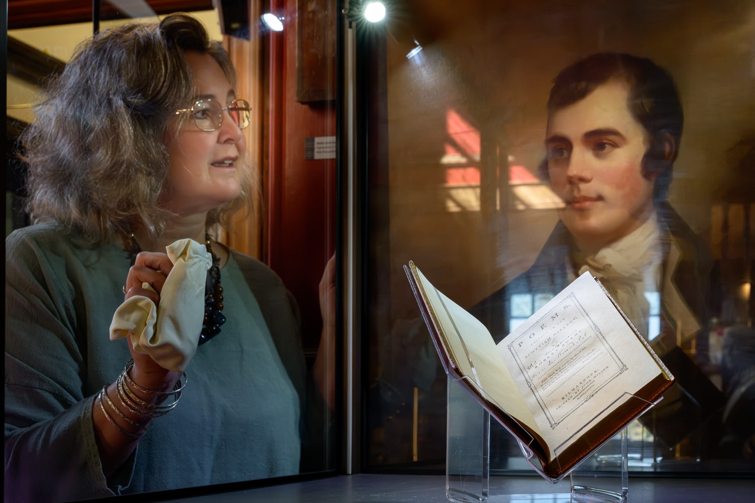 Kilmarnock Edition and a picture of Robert Burns in a glass case. A curator wiping the case.