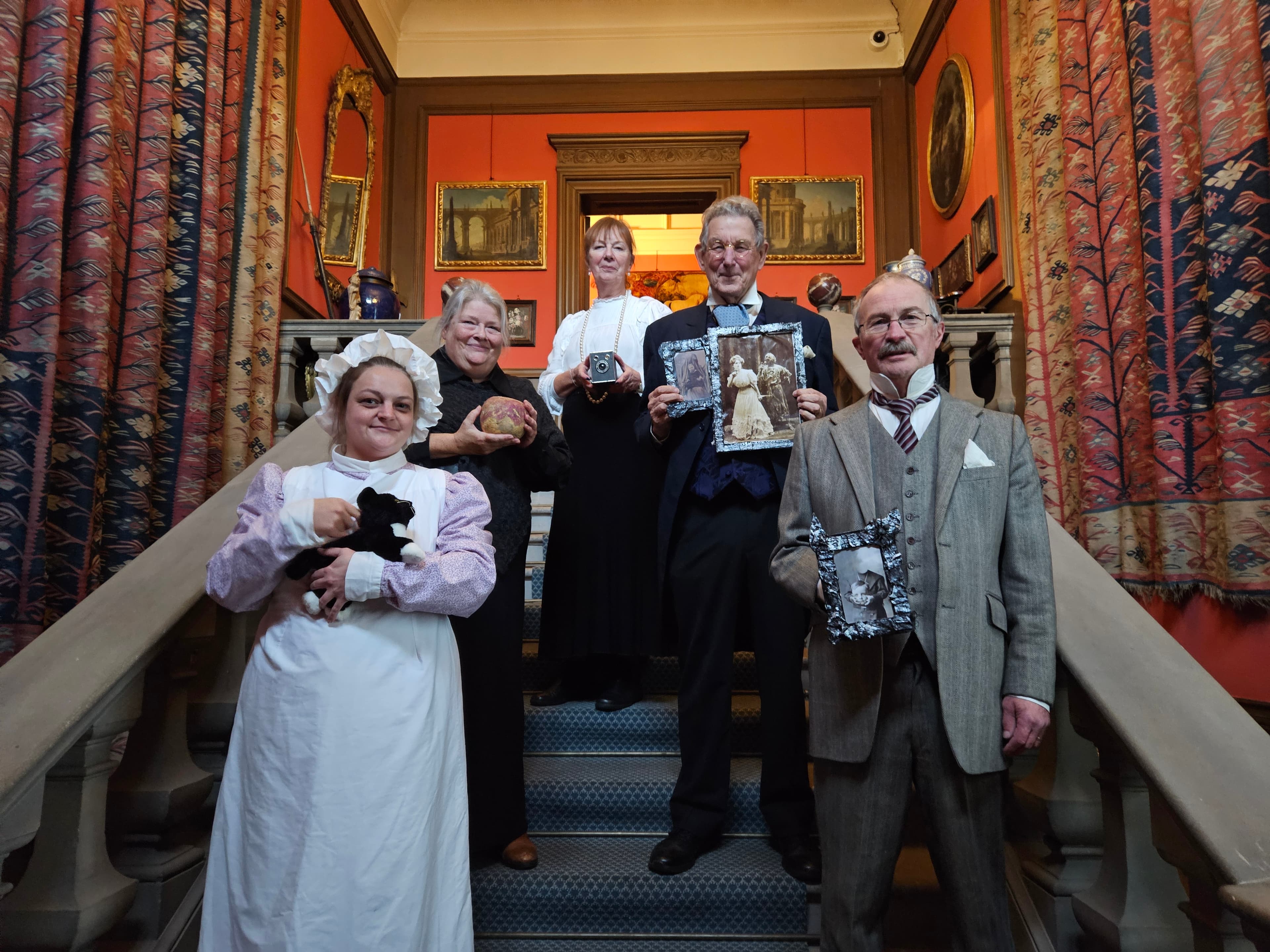 Five adults in edwardian period costume stand on a staircase, each holding an object, in an ornately decorated room with paintings, red walls, and patterned curtains.
