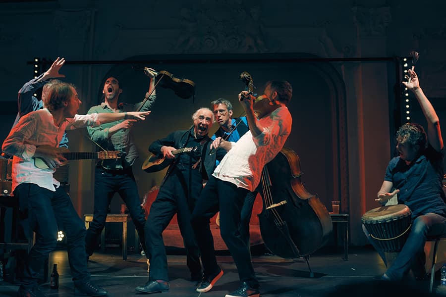 Image shows musicians on stage playing fiddles, guitars, and double bass with exaggerated expressions