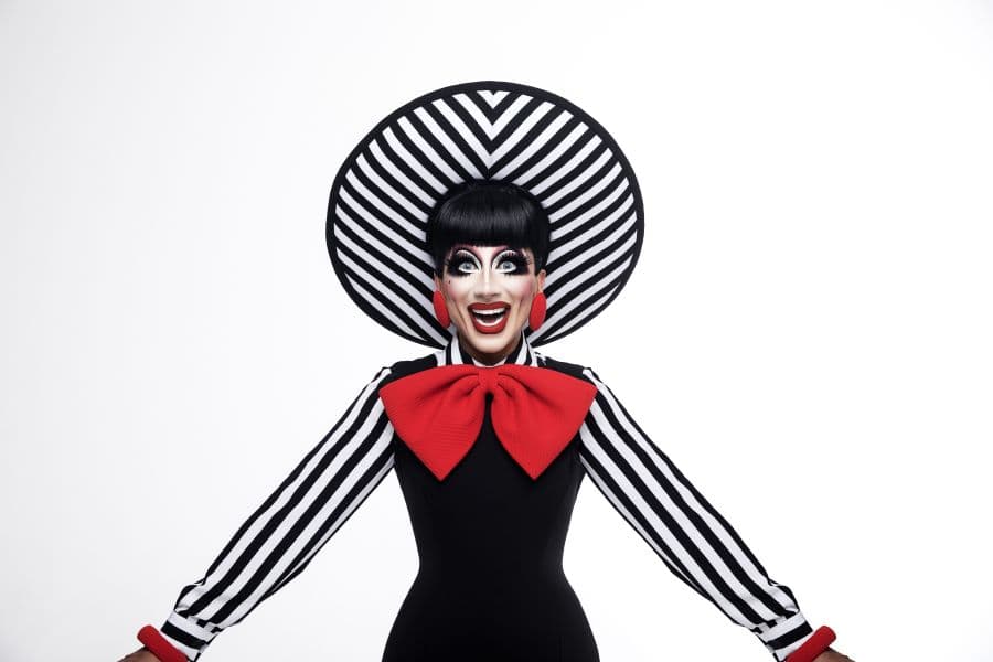 Image shows Bianca Del Rio smiling wearing black & white striped hat, black dress, and large red bow