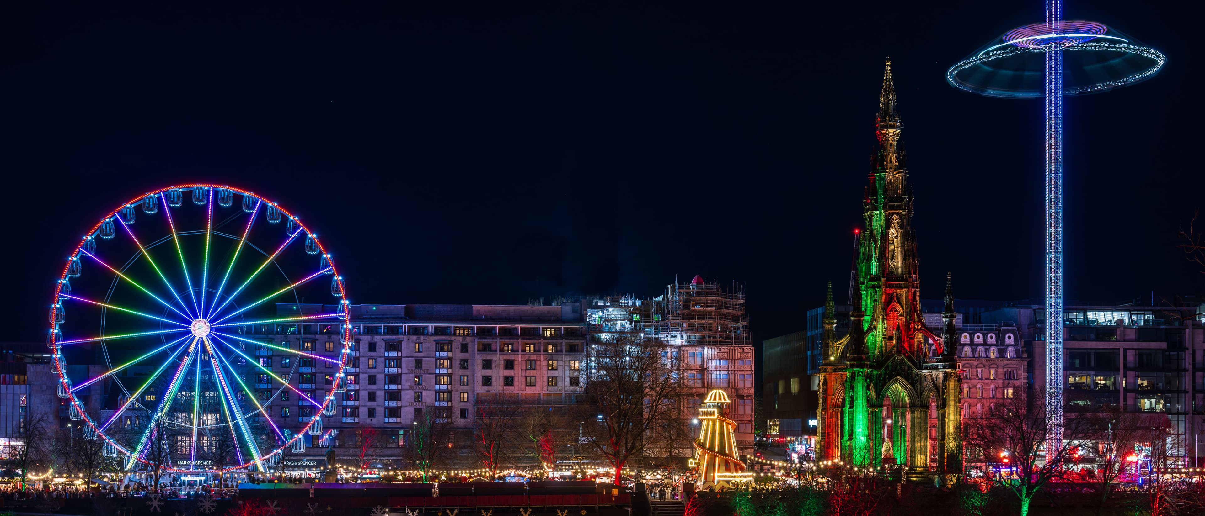 A dark night time photo showing coloured Christmas lights and Christmas fair rides, including the Scott Monument all lit up in green and red lights