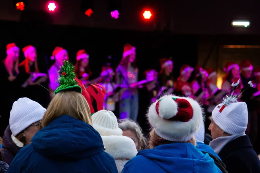 A crowd wearing winter hats and festive headbands watches a choir perform on stage under stage lights.