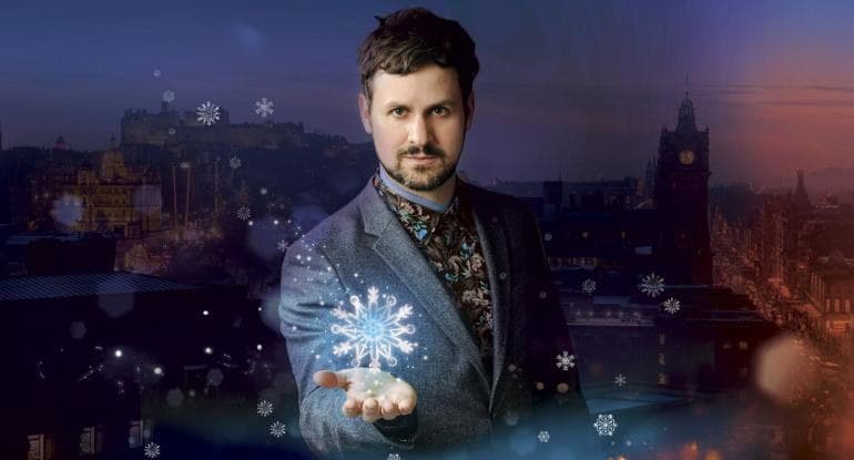 Image of Magician Kevin Quantum wearing grey suit and elobarate waistcoat. Ne is holding his hand outstretched with a snowflake on it.