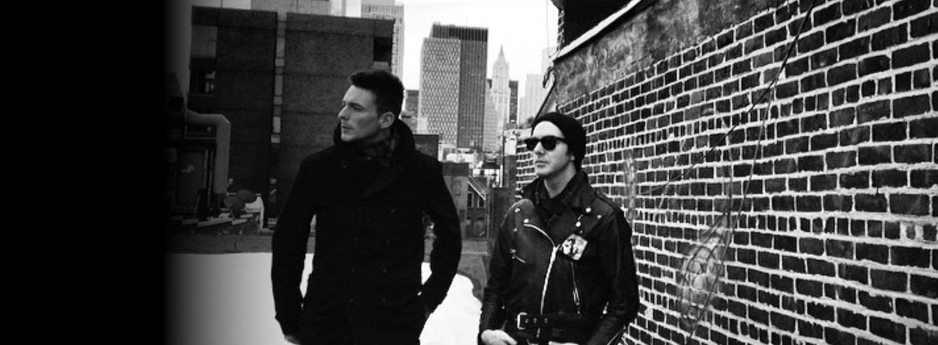 Two members of the rock band, Glasvegas, stand side by side in front of a textured brick wall, dressed casually.