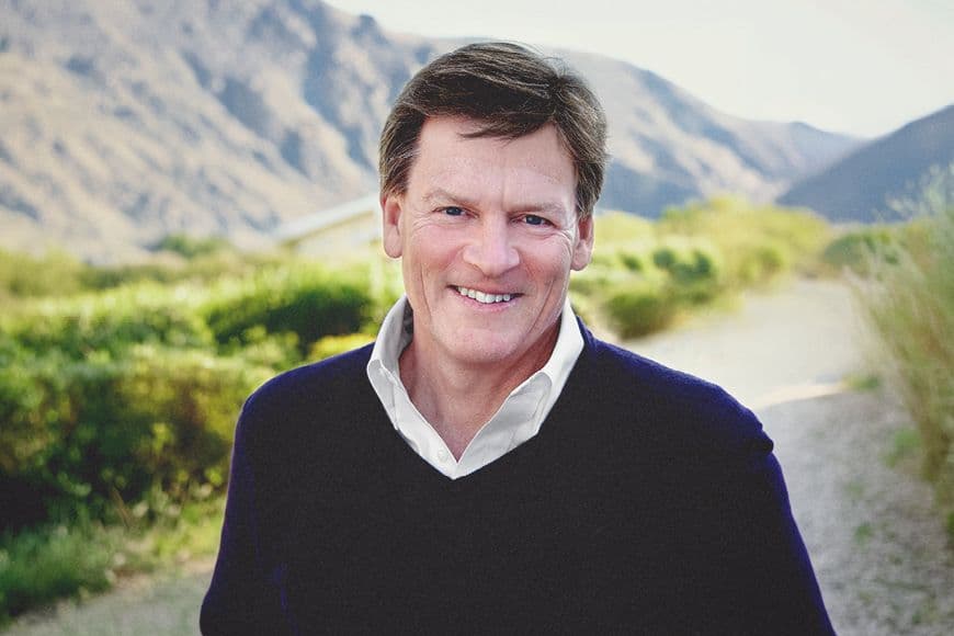Michael Lewis, smiling, wearing black v neck jumper and white shirt with green hills behind him.