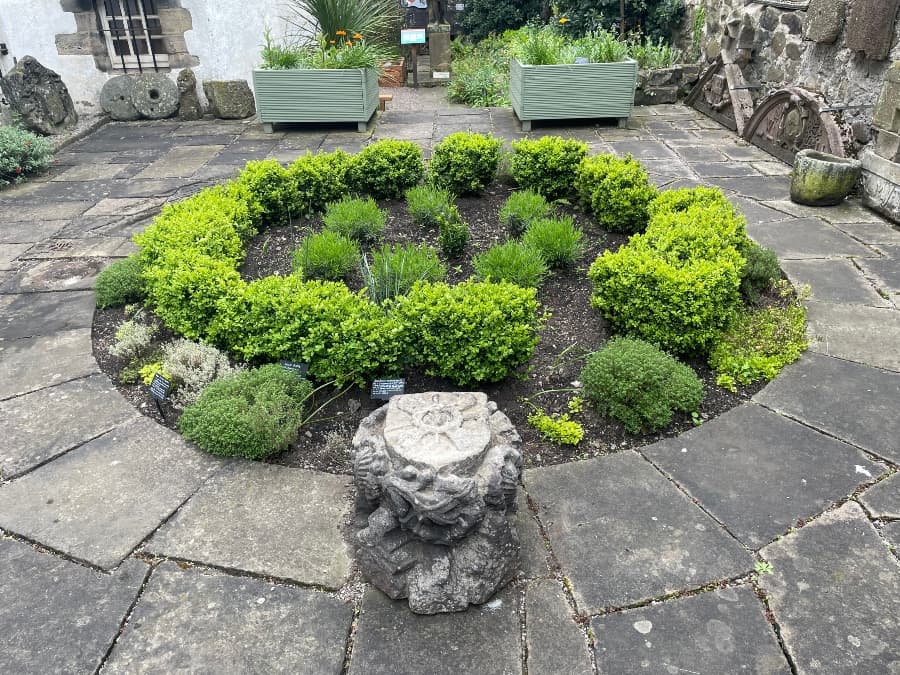 A circular garden bed edged with green shrubs within the courtyard at the Museum of Edinburgh. A carved 'passion stone' is in the foreground.