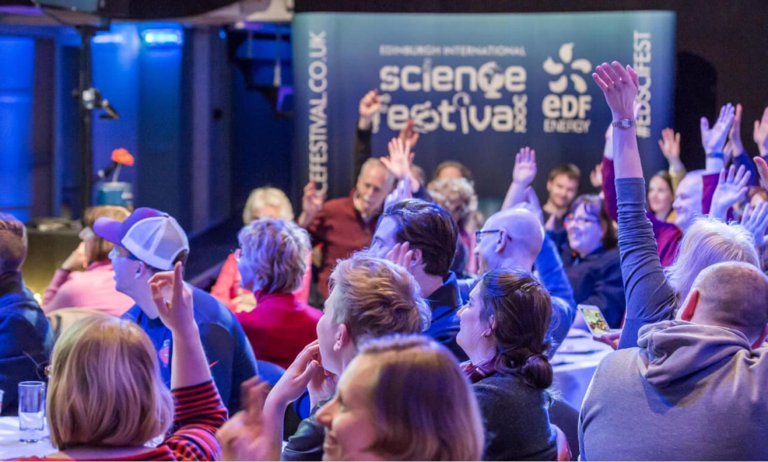 An audience of children and adults at a Science Festival raising their hands in answer to a question. 