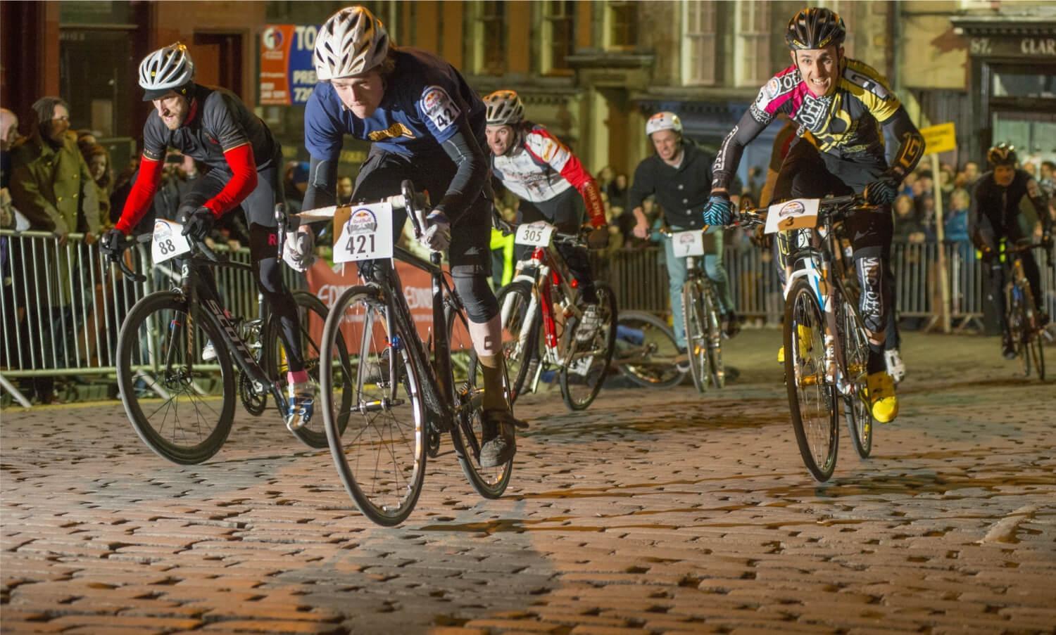 Five participants of Red Bull Hill Chasers event cycling up a cobbled street in Edinburgh with spectators on either side of the road behind metal barriers.